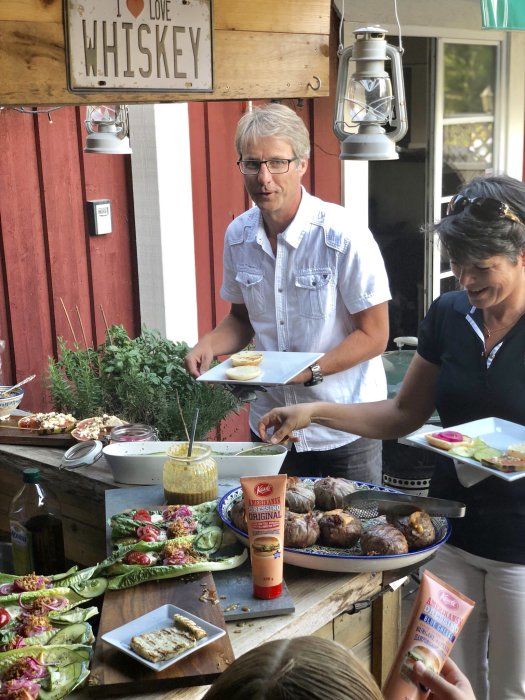 Personer förbereder måltid utomhus med baconinlindade burgare och tillbehör på serveringsbord.