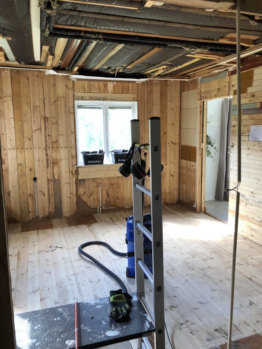 Ett delvis rivet rum med synliga isolering och ledningar i taket samt en stege och byggmaterial.