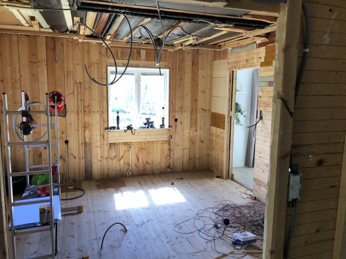 Ett rums interiör under renovering med nakna väggar och exponerade takbjälkar, kablar på golvet och en stege.