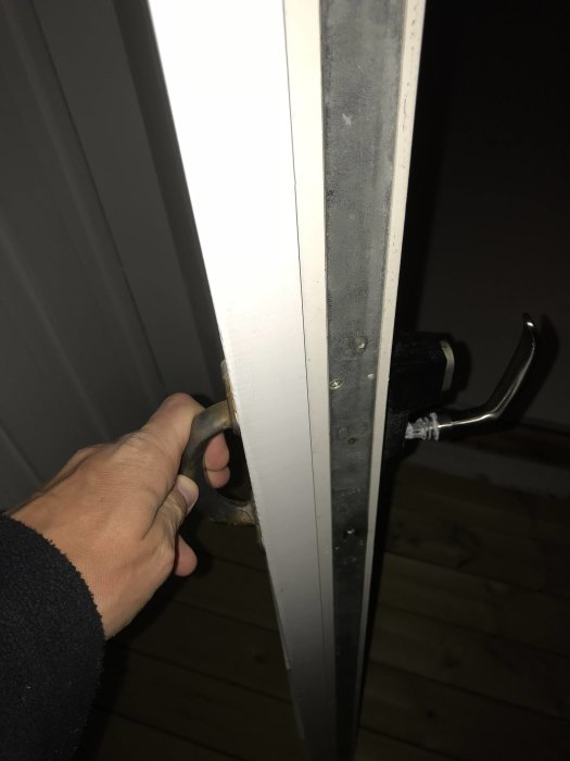 Hand håller ett defekt dörrhandtag med fastnitade skruvar synliga på dörrkanten.
