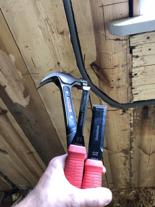 En hand som håller en hammare och en avbitartång framför elinstallationer på en vägg under renovering.