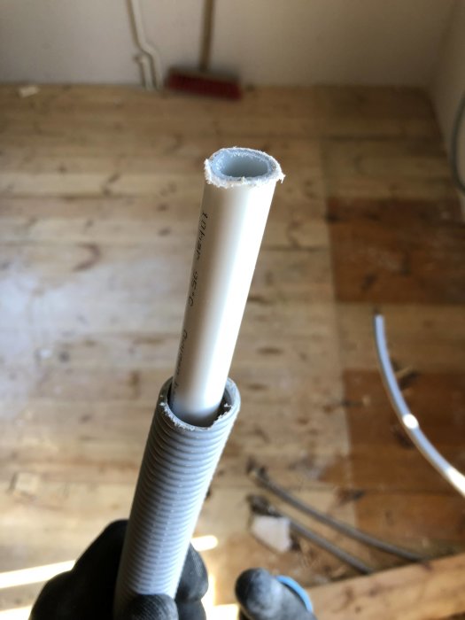 Närbild på en hand som håller i en alu-PEX rörände inuti ett flexibelt rörs skydd, på ett trägolv i bakgrunden.