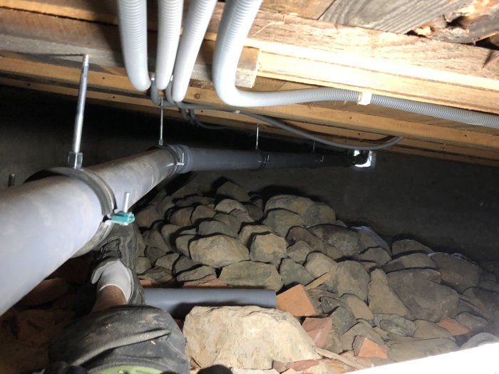 Avloppsrör installerade i krypgrund med stenig bas, samt synliga vattenrör och stöttor under en träbjälklag.
