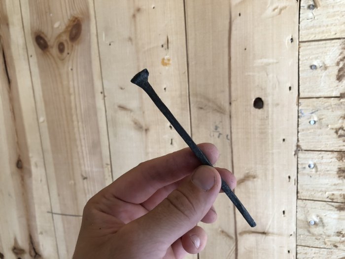 Hand håller en gammal rättad spik framför en trävägg.