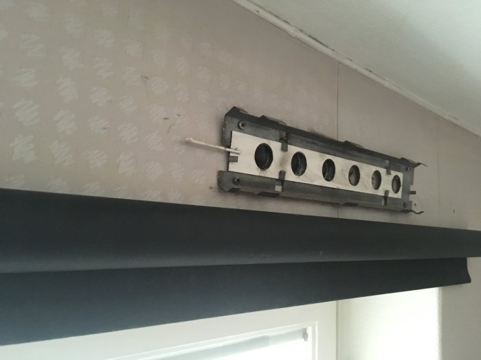 Veggen med flere runde hull og en metalldel over et vindu eller en åpning, sannsynligvis relatert til ventilasjon eller inspeksjon.