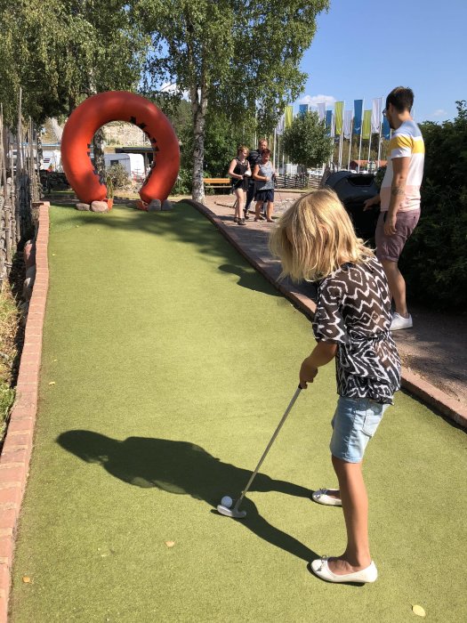 Barn spelar minigolf på en bana med en stor röd skulptur i bakgrunden under en klarblå himmel.