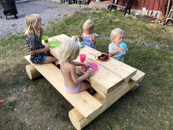 Barn sitter vid hemmagjort träbord utomhus och äter, med byggmaterial i bakgrunden.