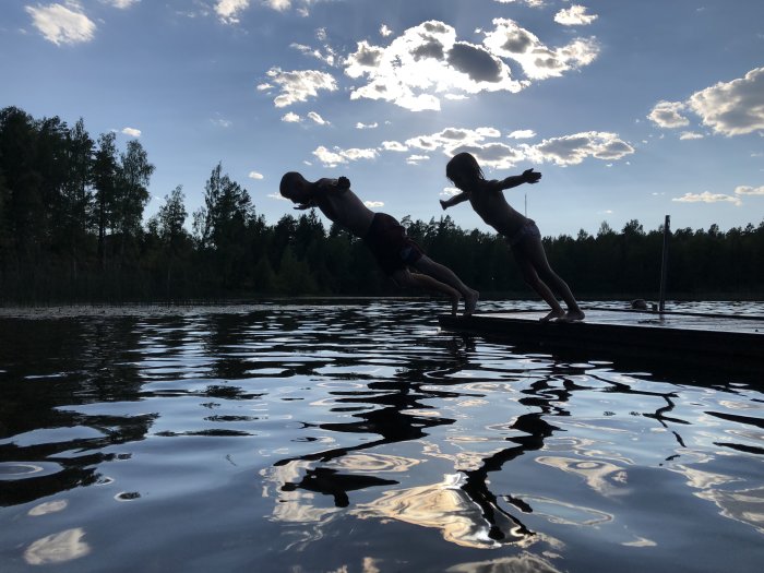 Två barn hoppar från en brygga i en sjö med skog i bakgrunden och kvällssol genom moln.