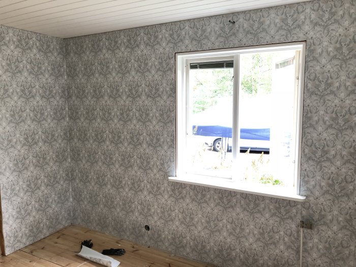 Nyligen tapetserad vägg i rum med blommigt mönster, synligt fönster och golv som väntar på ytterligare arbete.
