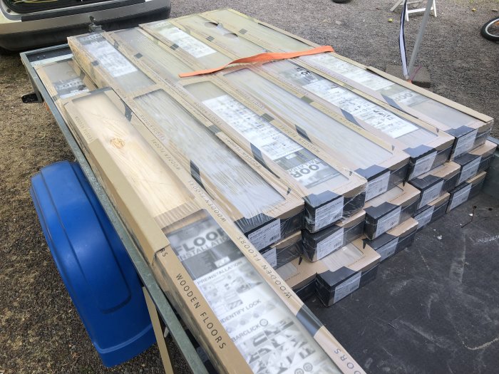 Förpackade 1-stav ljusgrå parkettgolv staplade utomhus nära en bil och byggmaterial.