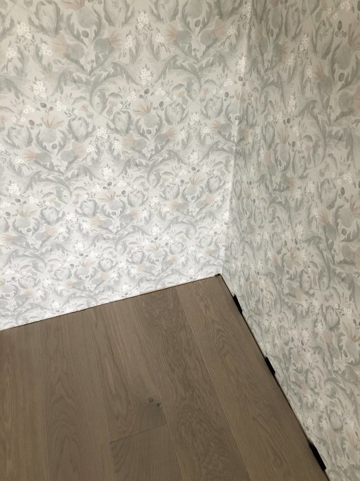 Ett hörn av ett rum med ljusgrått 1-stavs parkettgolv och vägg med blommigt tapetmönster.