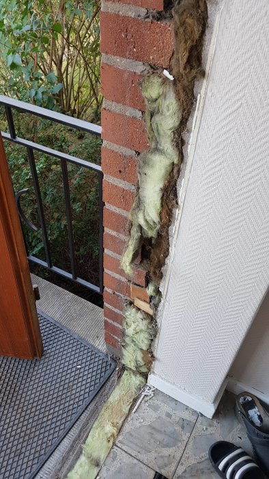 Öppet utrymme vid en ytterdörruttagning med exponerat isoleringsmaterial och träbitar mot en tegelfasad.