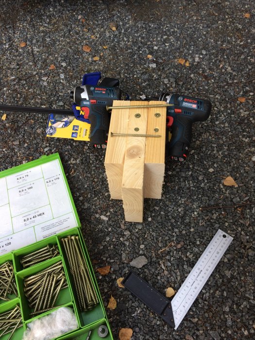 Bosch 12V-skruvdragare bredvid träbit med inskruvade skruvar, skruvlåda och tumstock på grusunderlag.