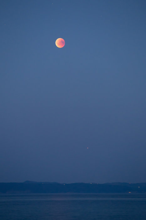 Blodmåne och Mars ovanför en belysningsprydd kustlinje vid Vätterstranden i aftonljus.