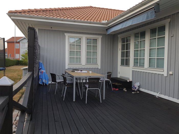 Bild av husets hörna med mörk trädäck, bord och stolar, samt ett litet fönster under takutsprånget.