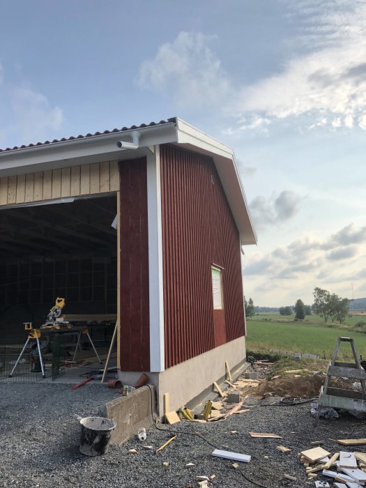 Färdigställd röd gavel på lada med byggmaterial och stege framför, under blå himmel.