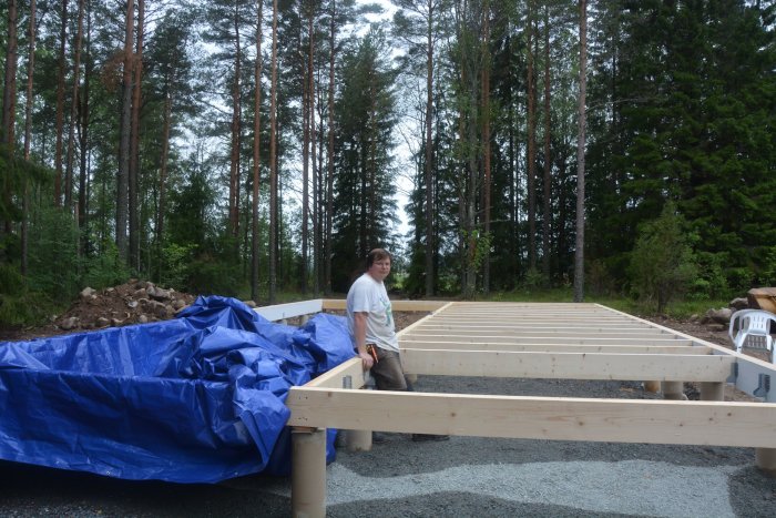 Nybyggt träbjälklag för husgrund med skyddande blå presenning och person i arbetskläder, i skogsmiljö.