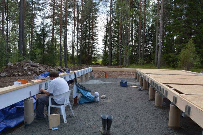 En person bygger på ett nytt bjälklag för ett hus, omgivet av en skogsmiljö och byggmaterial på gruset.