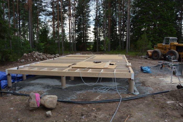 Nyttbjälklag för hus på betongpelare i skogsmiljö med verktyg och byggfordon i bakgrunden.