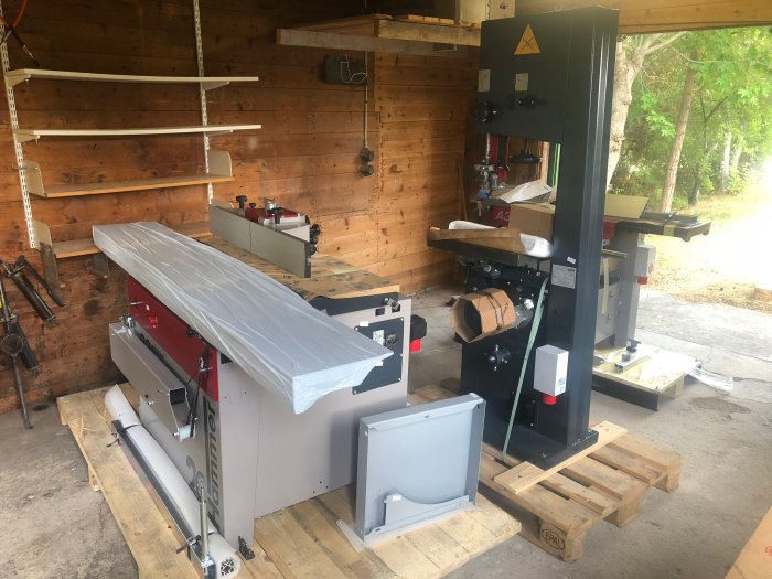Ny träbearbetningsutrustning i ett garage, inklusive en bandsåg och en tjocklekshyvel på pallar.