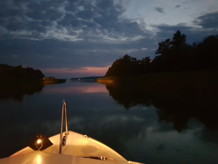 Nattbild från båt på Bälstaviken med lugnt vatten, silhuetter av träd och ljus från Prippsbron i fjärran.