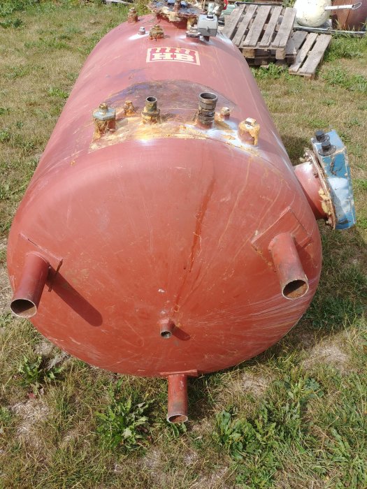 Stor röd tank på ca 110cm i diameter med tömningskran och rostiga fästelement på gräs.