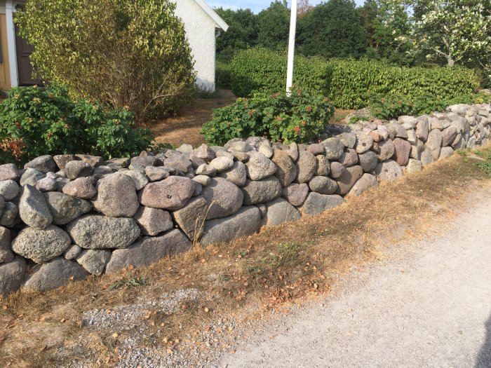 Kallmurad stenmur bestående av kluvna stenar vid en väg, med grönska och en vit byggnad i bakgrunden.