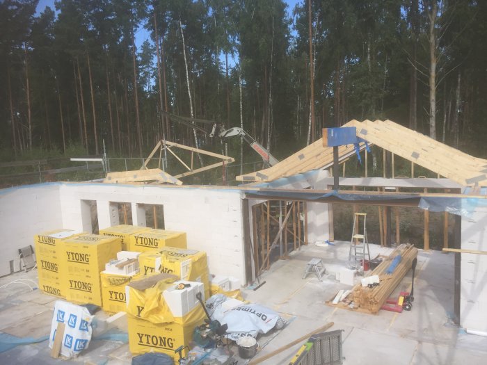 Byggplats med nästan färdigställda takstolar på en husgrund och byggmaterial framför skogsbakgrund.