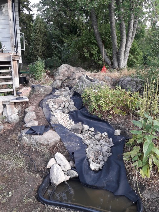 Pågående byggprojekt med stenbädd och svart duk, ställbar pump och trädgård i bakgrunden.