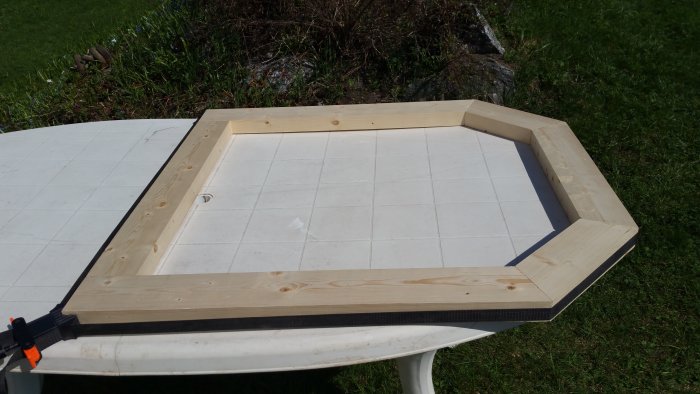 Omonterad ram till ett altanbord i furu, placerad på ett runt vitt bord ute i solen.