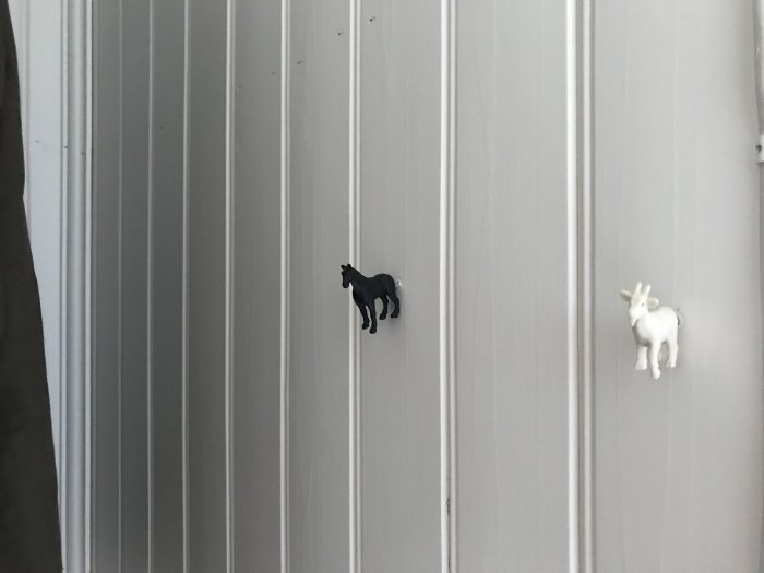 Plastdjurskrokar på vit panelvägg, svart häst till vänster och vit get till höger.