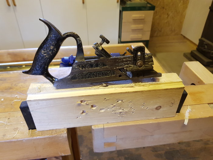 Antik falshyvel med brutet framstycke placerad på träbit, omgiven av hyvelspån.