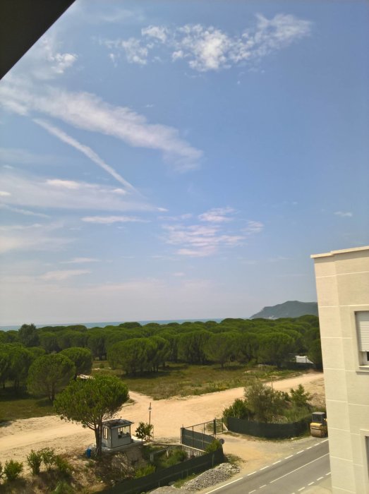 Utsikt från ett fönster över en väg, träd och berg i fjärran under en delvis molnig himmel.