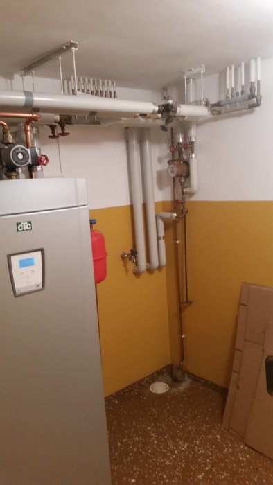 Värmepumpsinstallation med synliga röranslutningar, cirkulationspump och ventilhus i ett förrådsutrymme.