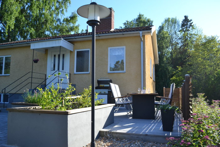 Renoverad trädgård med trall, odlingslåda, matplats och marksten framför gult hus.