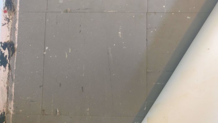 Hörn av källargolv med upplyft plastmatta som avslöjar gamla hårda plastplattor och svart lim på väggen.