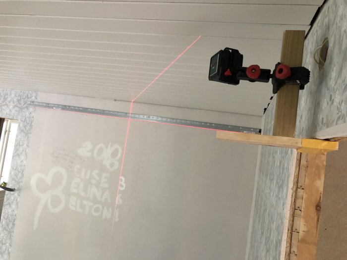 Laseravståndsmätare monterad på träplank projicerar en röd linje för exakt mätning i renoveringsprojekt.