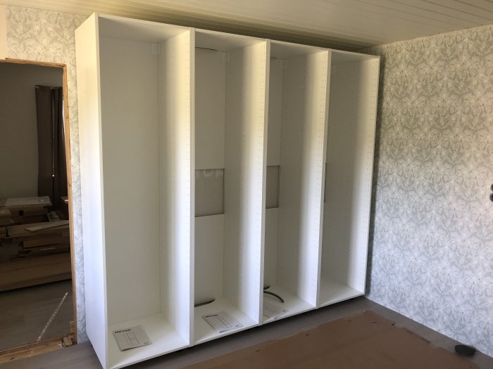 Omonterade IKEA-skåpstommar i vitt, placerade i ett rum med tapetserade väggar under pågående renoveringsprojekt.