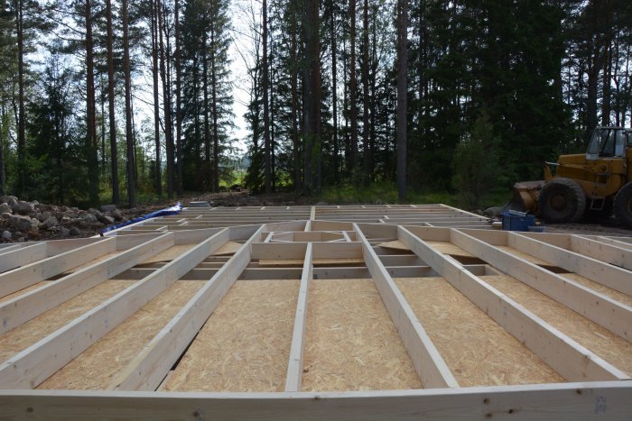 Nybyggd trägolvstomme för hus på en byggarbetsplats med skog i bakgrunden och en gult hjullastare till höger.