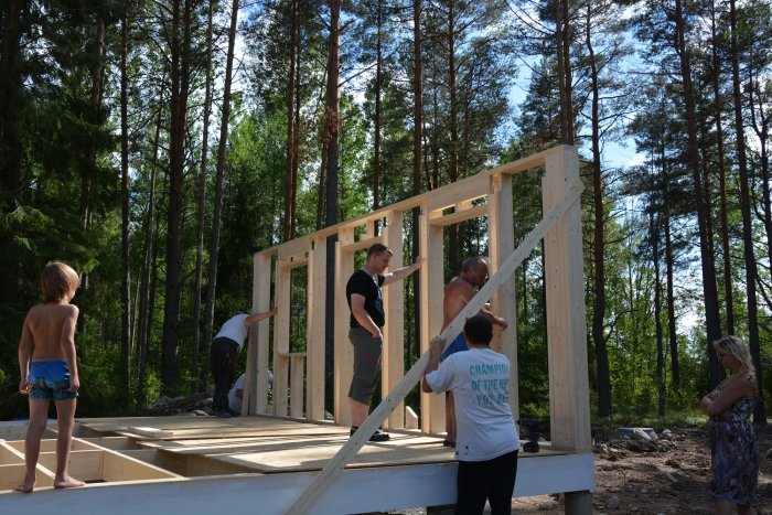 Familj hjälper till med att resa en hög trävägg på en husgrund i skogsmiljö.