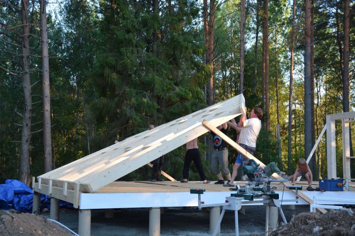 Fem personer reser en hög trävägg på en byggplats i skogsmiljö.