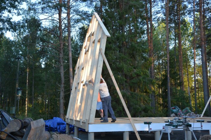 Två personer reser en stor trävägg på ett husbygge i en skogsmiljö under kvällssol.