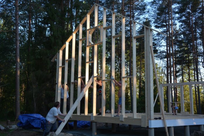 Fem familjemedlemmar reser en hög trägavelvägg på ett husbygge bland träd.