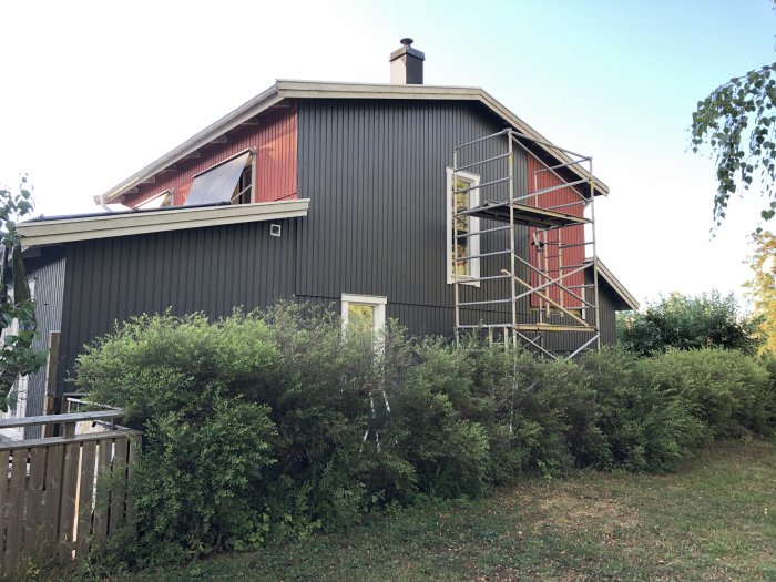 Ett hus under renovering med nypanelade fönstersmygar, ställning vid gavelfönstret, omgiven av grönska.