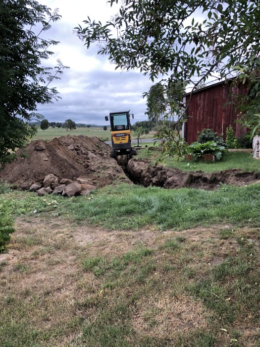 Minigrävare vid öppnad grävd ränna i trädgård nära röd lada, omgiven av högar med grus och jord.