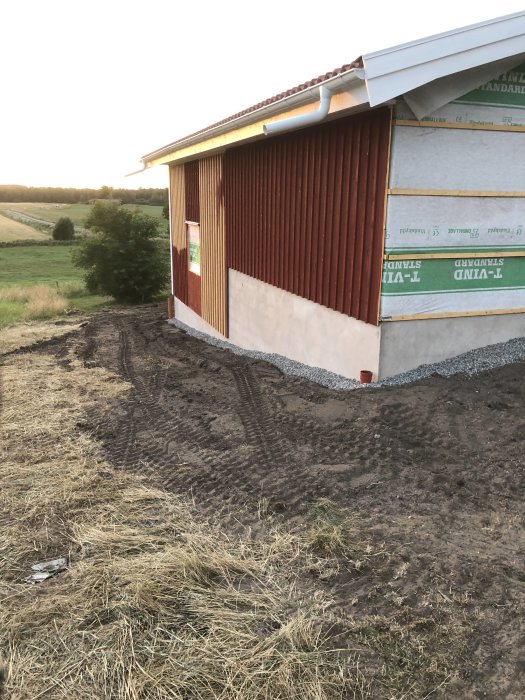 Nygrusat område vid röd garagebyggnad med isolering på väggen, spår av grävning syns och en del grönt gräs.