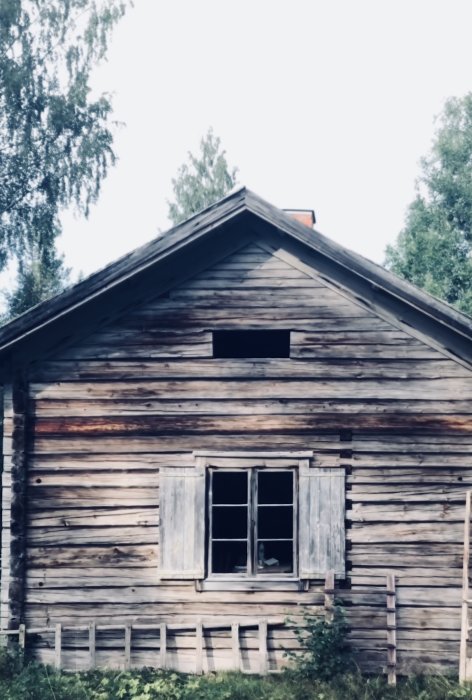 Gammalt timmerhus med rektangulärt och runt hål i vinden, förfallen fönsterlucka.