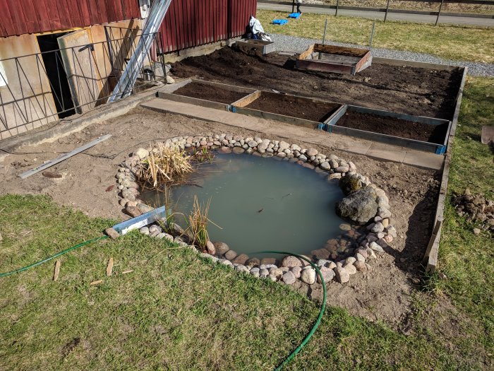 En liten damm omgiven av stenar i en trädgård under anläggning, med pågående arbete och odlingsbäddar i bakgrunden.