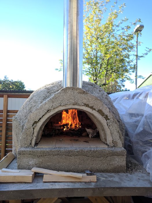 Ugnseld i hembyggd vedeldad pizzaugn av vermikulit med synlig rökkanal och vedträn framför.