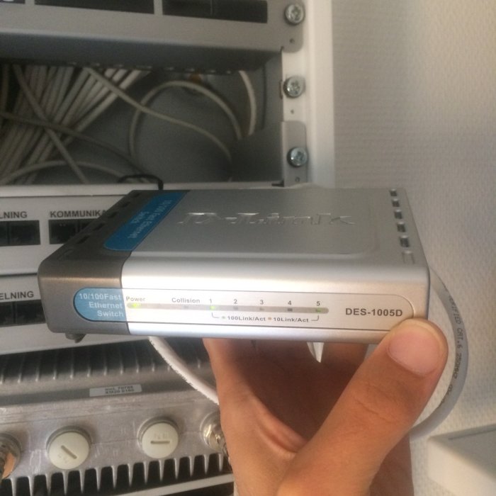 Hand håller en D-Link DES-1005D switch med synliga Ethernet-portar och kablar i skåpet bakom.